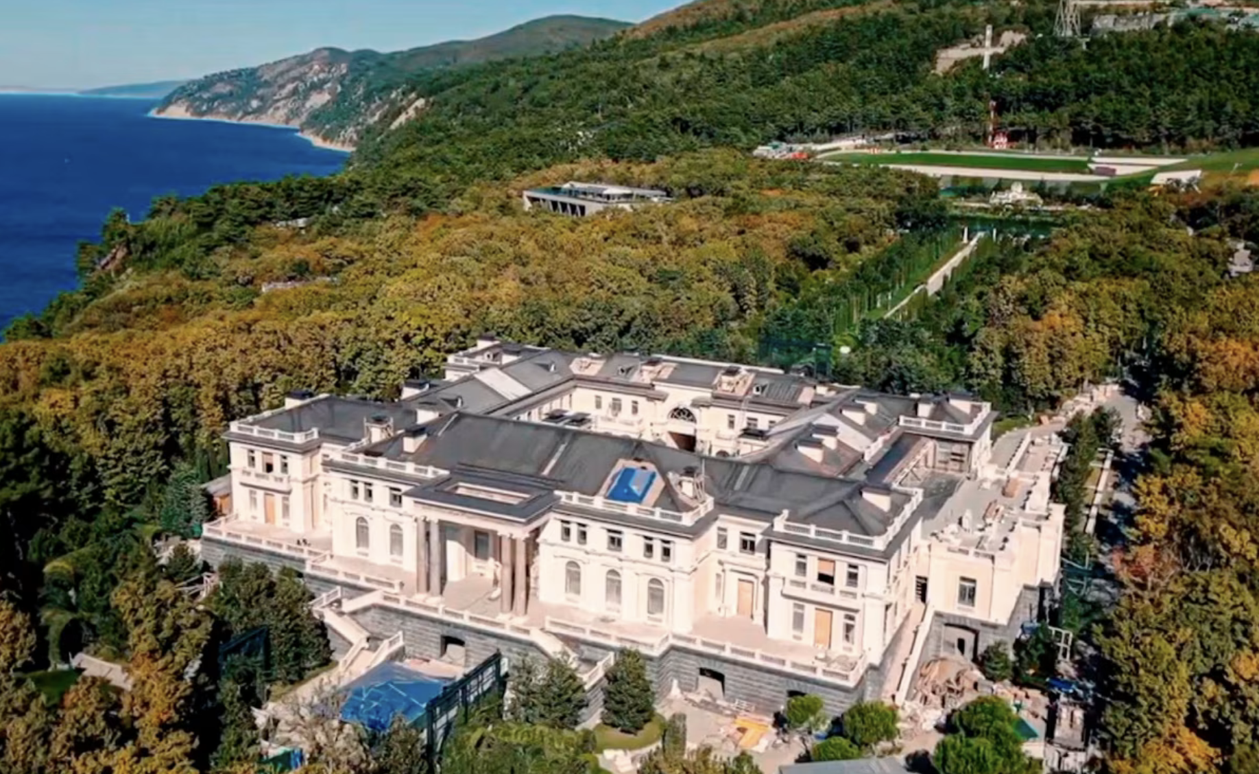An aerial view of a very large white building surrounded by trees, with the sea in the distance
