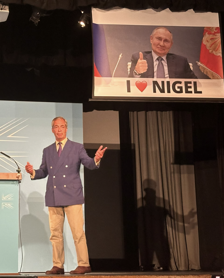 Nigel Farage wearing a blazer and gesturing onstage near signs that say "Reform U.K." and "Britain Needs Reform." People on either side of him are clapping.